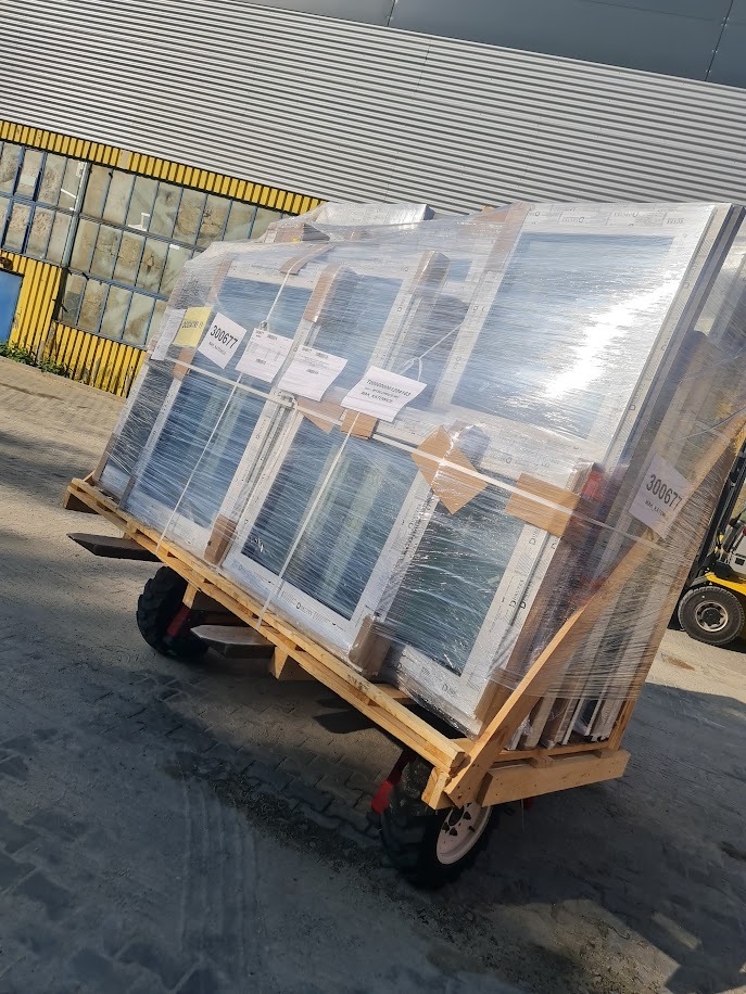 Paleta z oknami PCV, owinięta folią, przewożona na wózku transportowym na zewnątrz budynku o metalowej fasadzie. Widoczne etykiety na oknach.