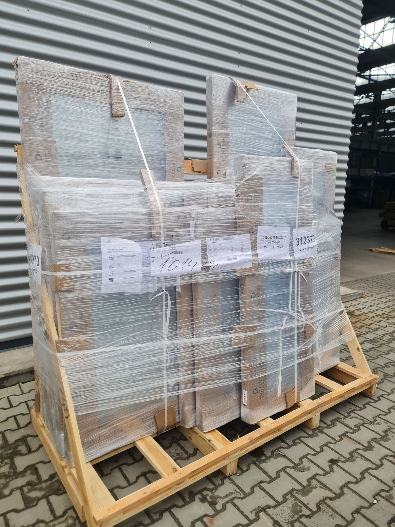 Paleta z zapakowanymi oknami lub drzwiami, zabezpieczonymi folią stretch i taśmami. Widoczne etykiety z numerami seryjnymi i nazwą producenta.