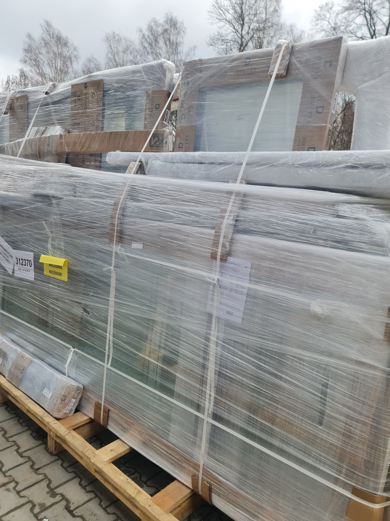Stos okien PCV marki Drutex, zabezpieczonych folią, na palecie w Siemianowicach Śląskich. Widoczna etykieta z akcesoriami i oznaczeniami producenta.