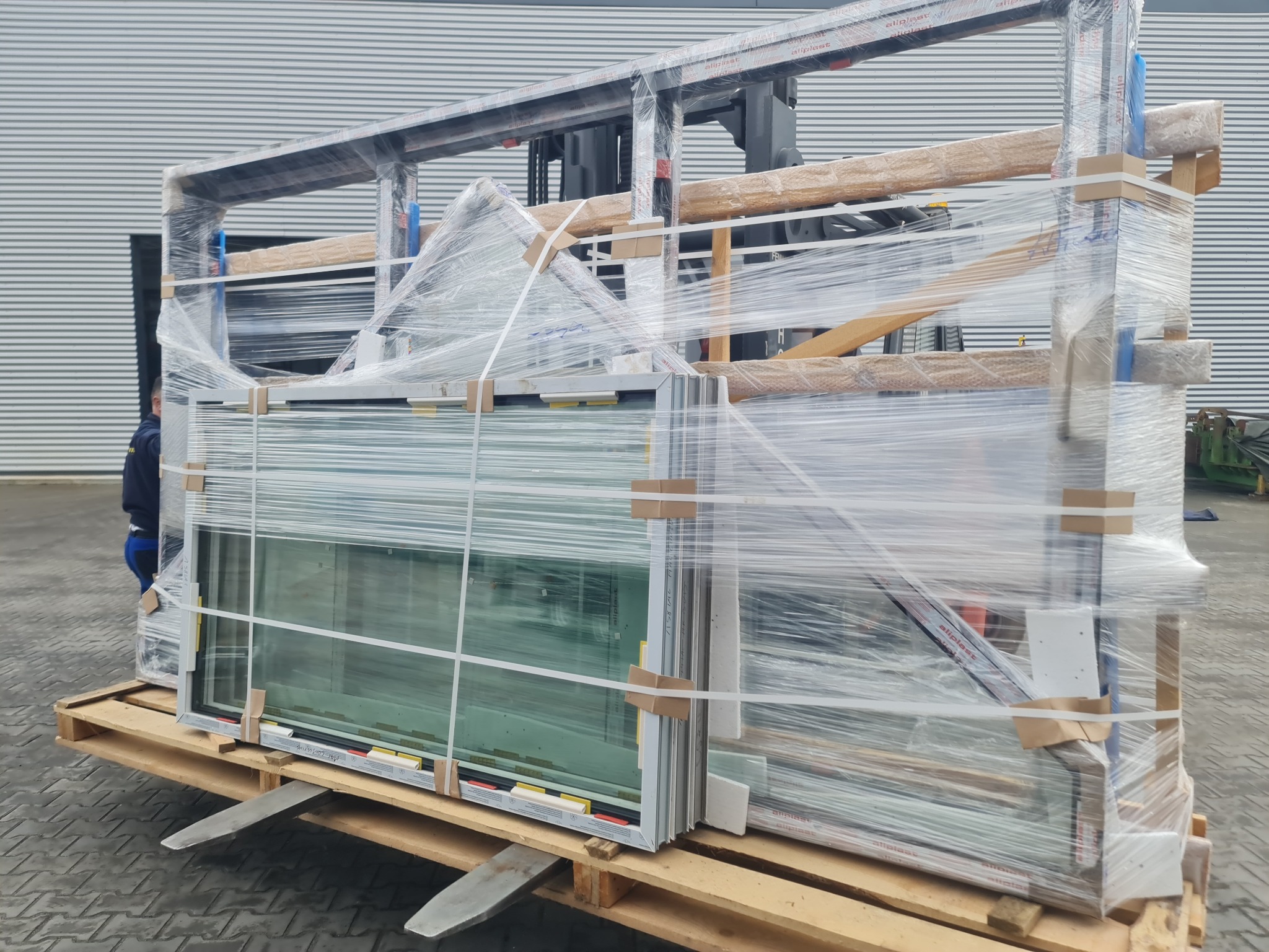 Paleta z zapakowanymi oknami, zabezpieczonymi folią, transportowana wózkiem widłowym na zewnątrz budynku z szarej blachy trapezowej.