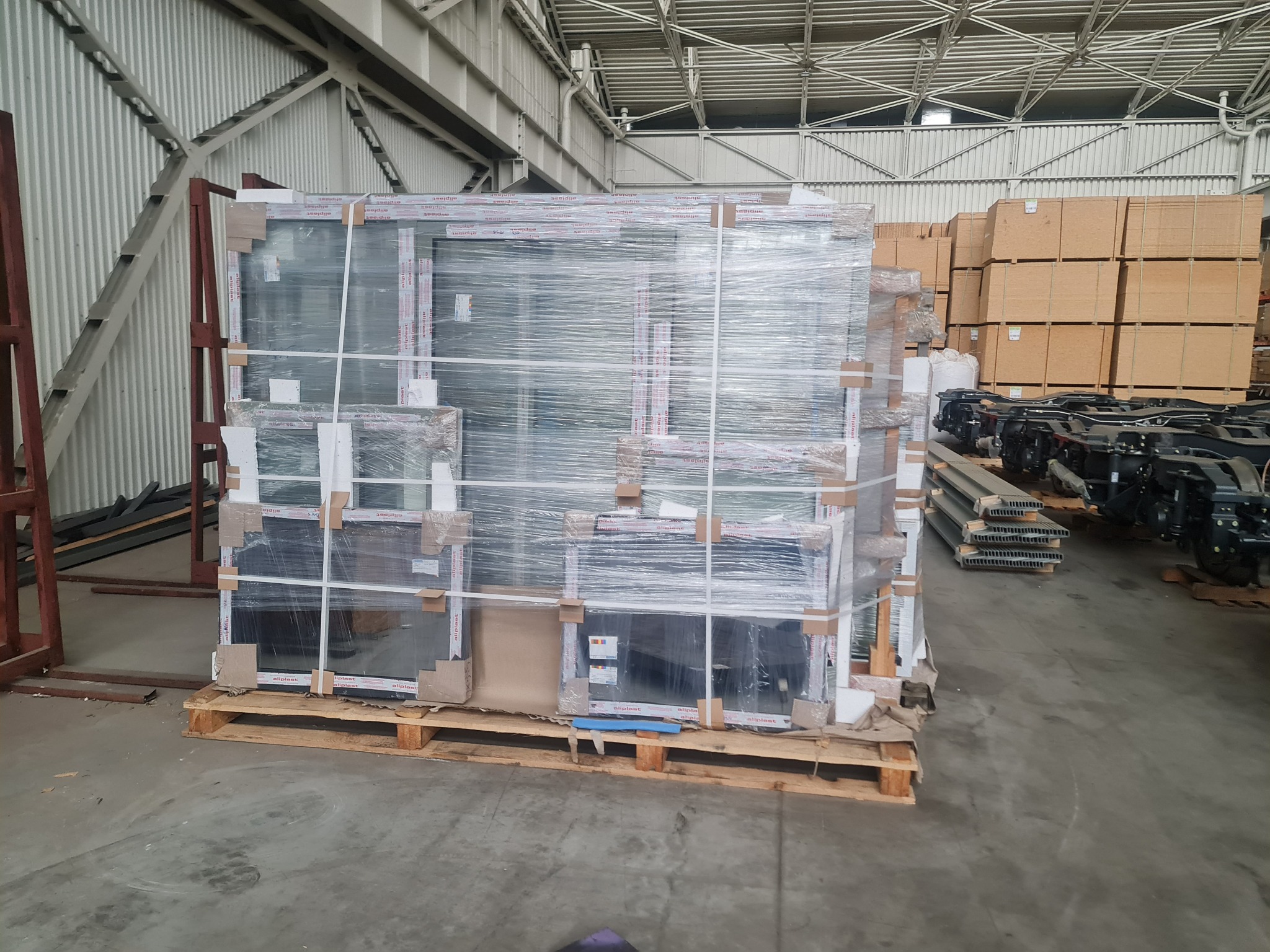 Paleta z zapakowanymi oknami PCV w magazynie, owinięta folią stretch, widoczne kartonowe zabezpieczenia narożników i etykiety producenta.