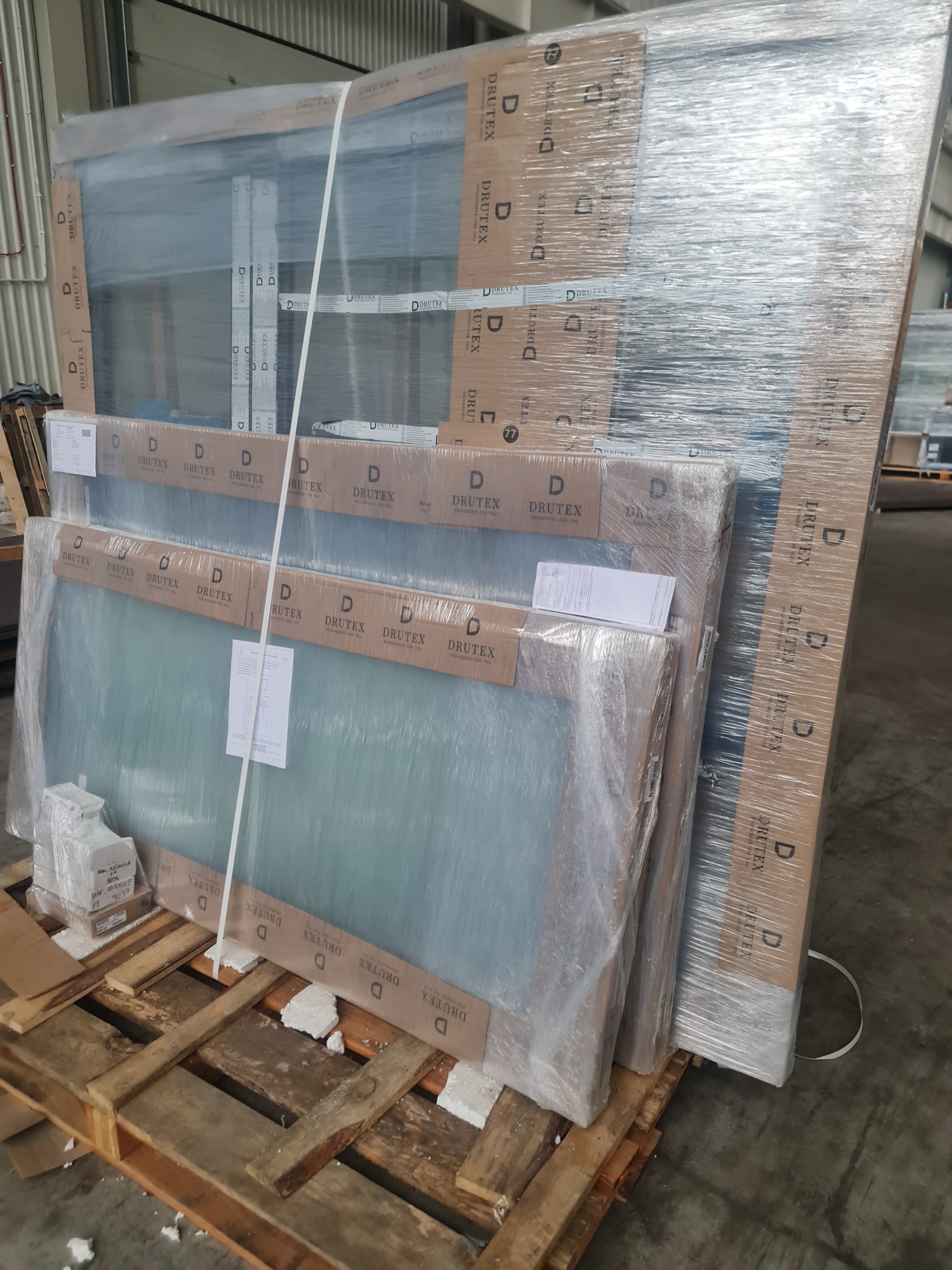 Paczka okien Drutex na palecie, zabezpieczona folią i kartonem z logo. Widoczne etykiety i dokumenty. Przygotowane do transportu w hali.