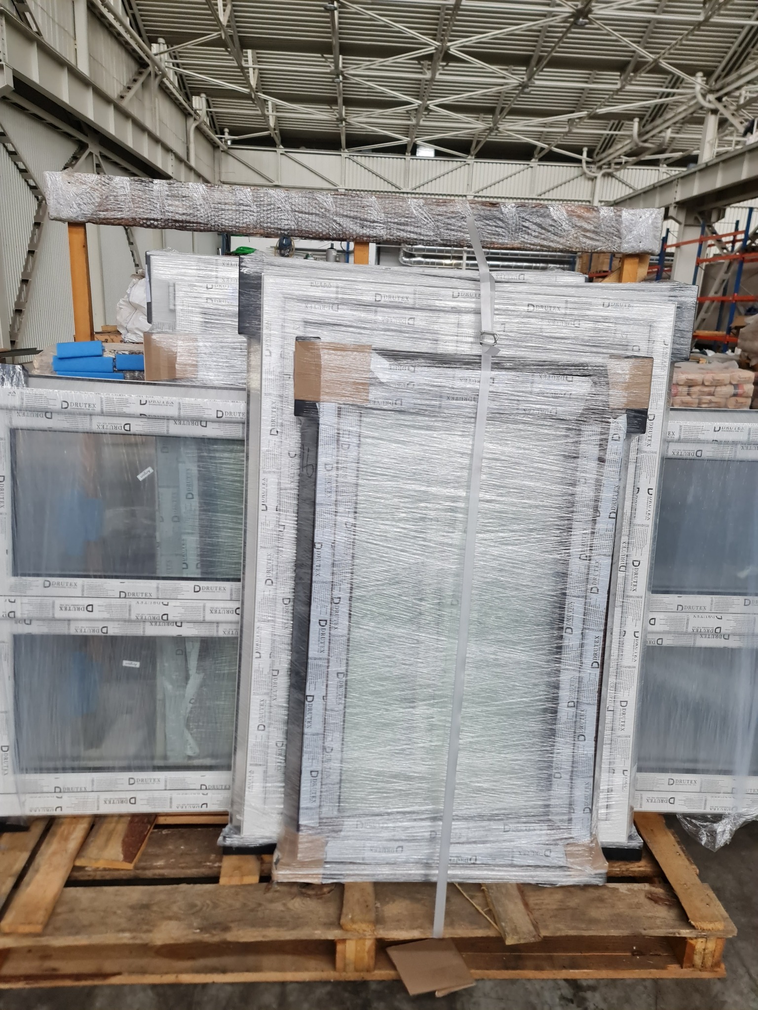 Sztapla zapakowanych okien marki Drutex na palecie w hali magazynowej. Okna owinięte folią stretch i zabezpieczone taśmą. Widoczne logo producenta na ramach.