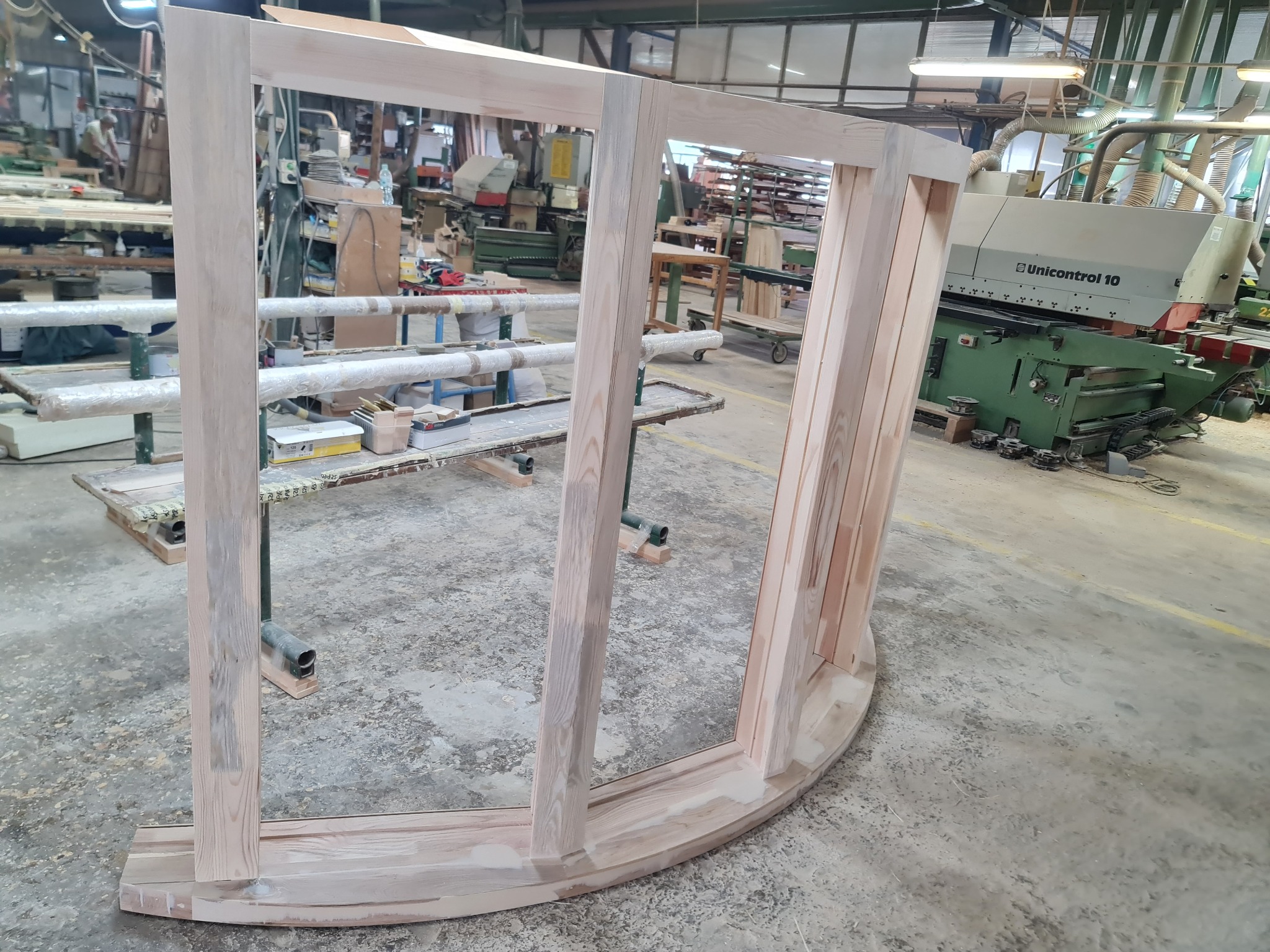 Drewniana rama okienna o zaokrąglonym kształcie, surowe drewno, widoczne słoje, w tle warsztat stolarski z maszynami i narzędziami, etap produkcji.