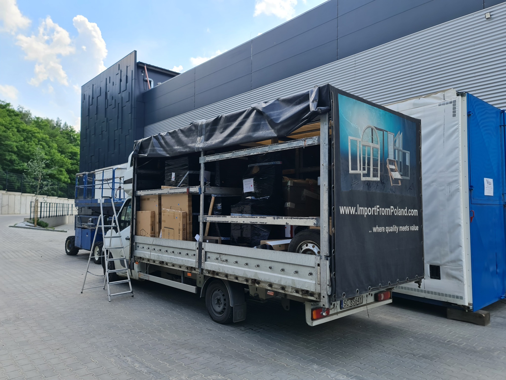 Ciężarówka z plandeką, załadowana oknami i materiałami budowlanymi, zaparkowana obok budynku z nowoczesną elewacją. Widoczny podnośnik i drabina.