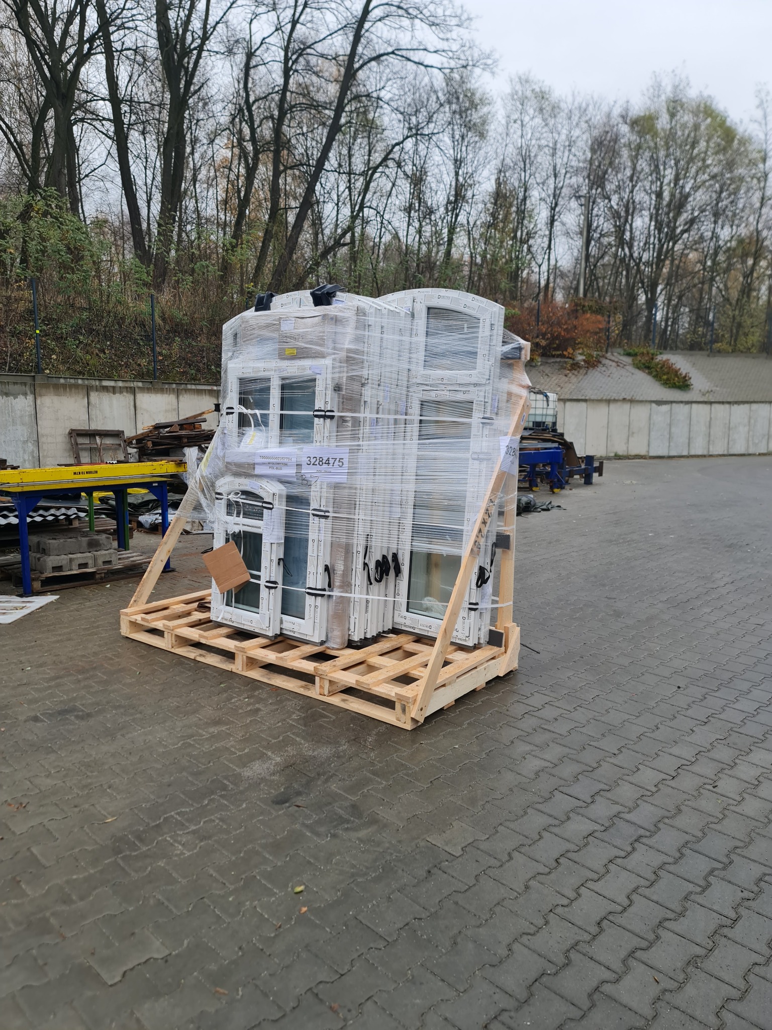 Paleta z zapakowanymi w folię białymi oknami PCV, oczekująca na montaż. Na oknach widoczna etykieta z numerem seryjnym. Tło: kostka brukowa, drzewa.