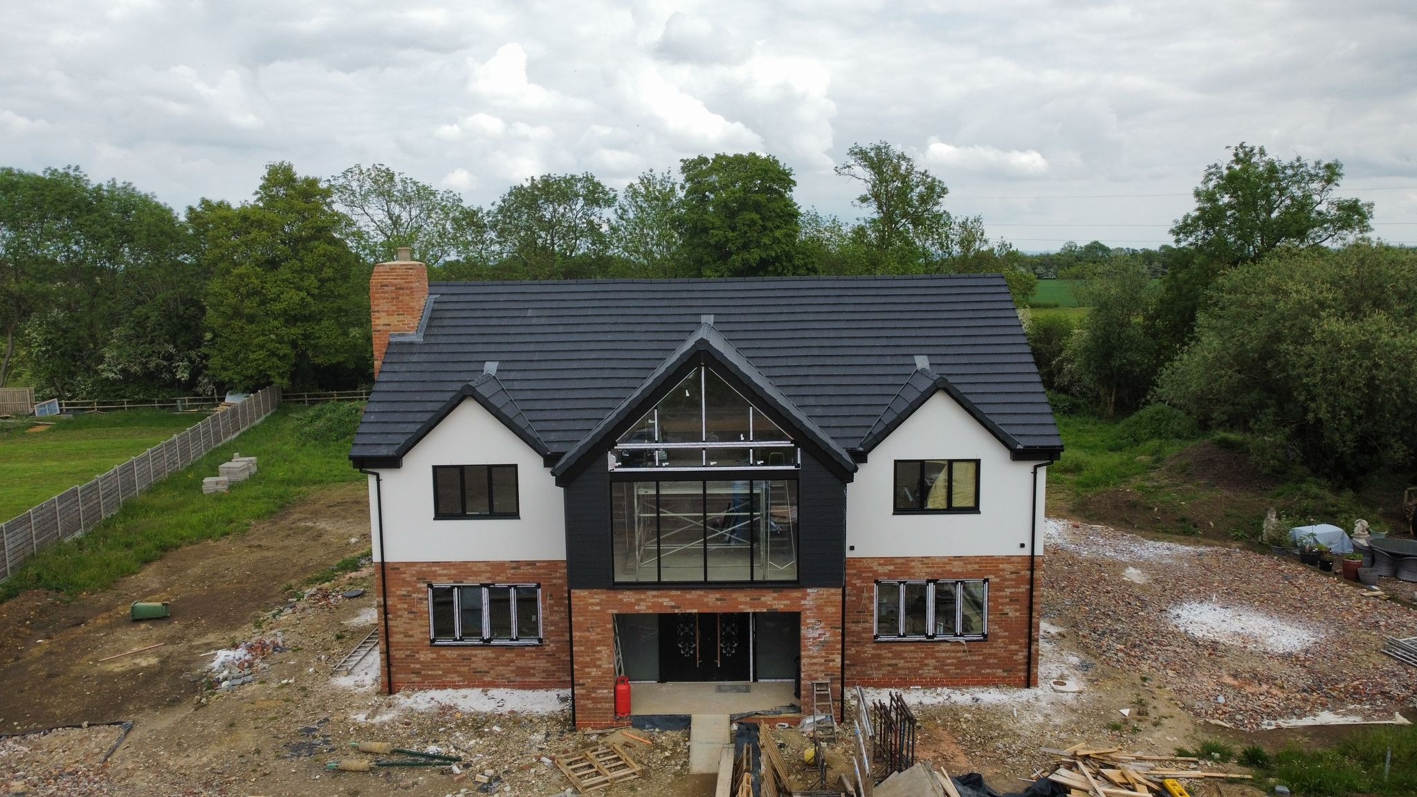 Dwukondygnacyjny dom z ciemną dachówką i dużymi oknami, w trakcie budowy. Widoczne rusztowania i materiały budowlane, tło stanowią drzewa i pochmurne niebo.