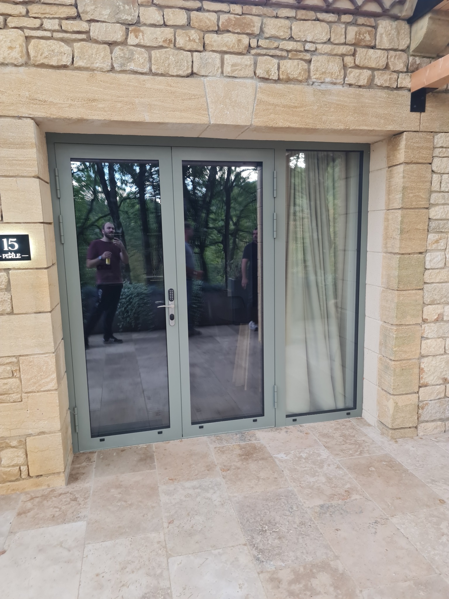 Szklane, trzyskrzydłowe drzwi tarasowe z szarymi ramami, wkomponowane w kamienną elewację budynku. W odbiciu widać zieleń ogrodu i sylwetki osób.