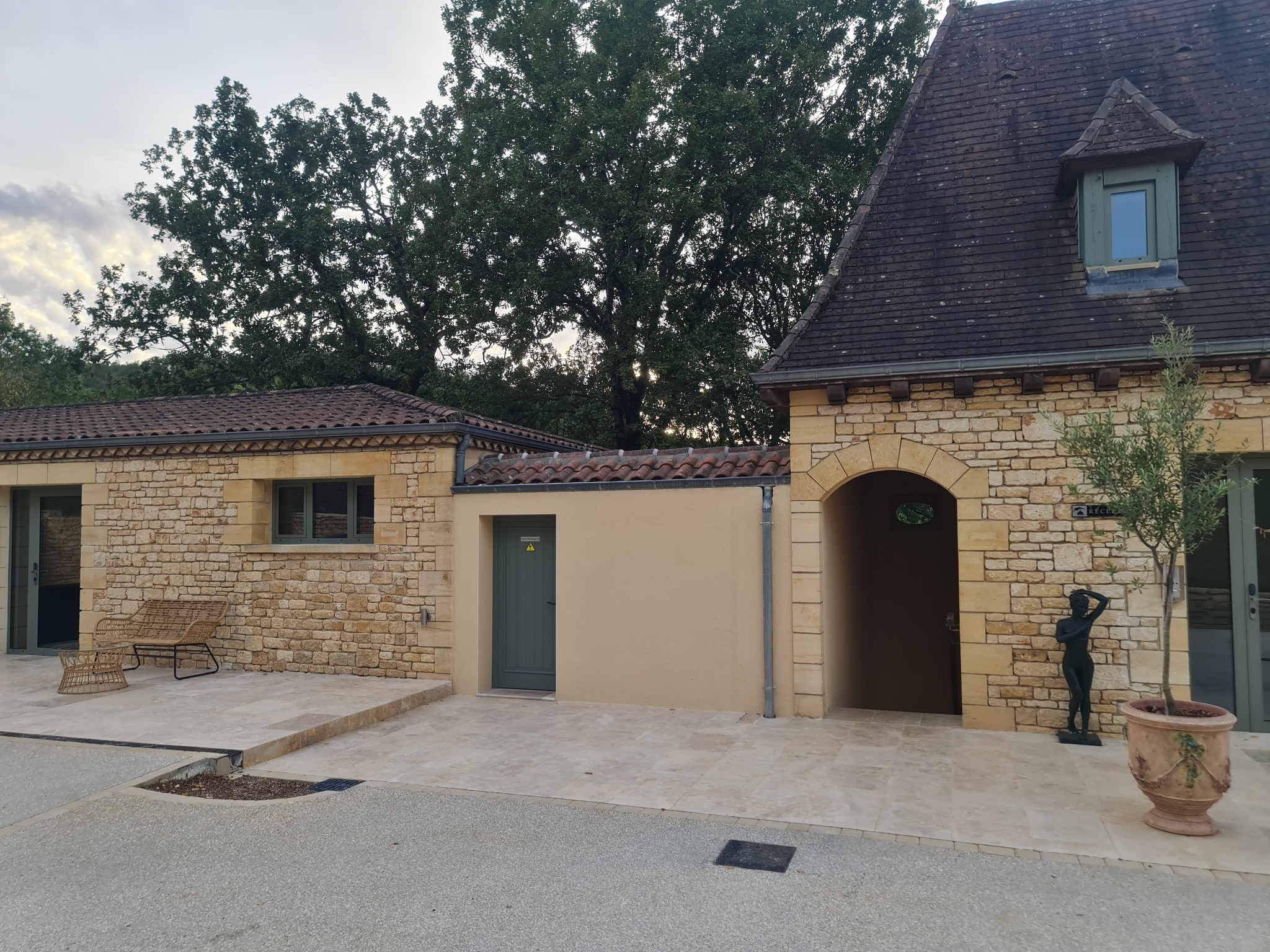 Elewacja budynku z kamienia z zielonymi drzwiami i oknami, brama łukowa, statua kobiety i drzewko w donicy. Tło stanowią drzewa.