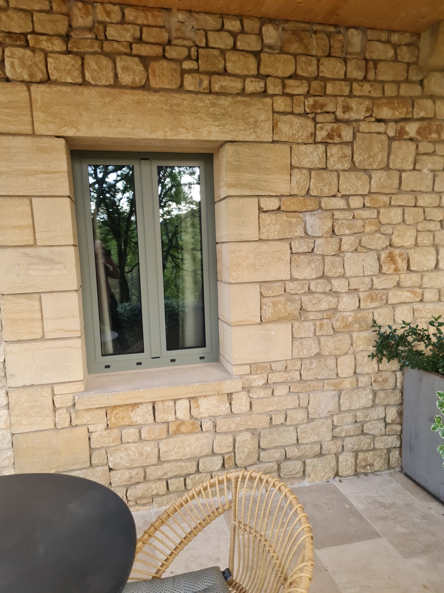 Szare okno w kamiennej ścianie, odbicie drzew w szybie. Elementy wystroju tarasu: stolik, krzesło z rattanu i donica z roślinami.