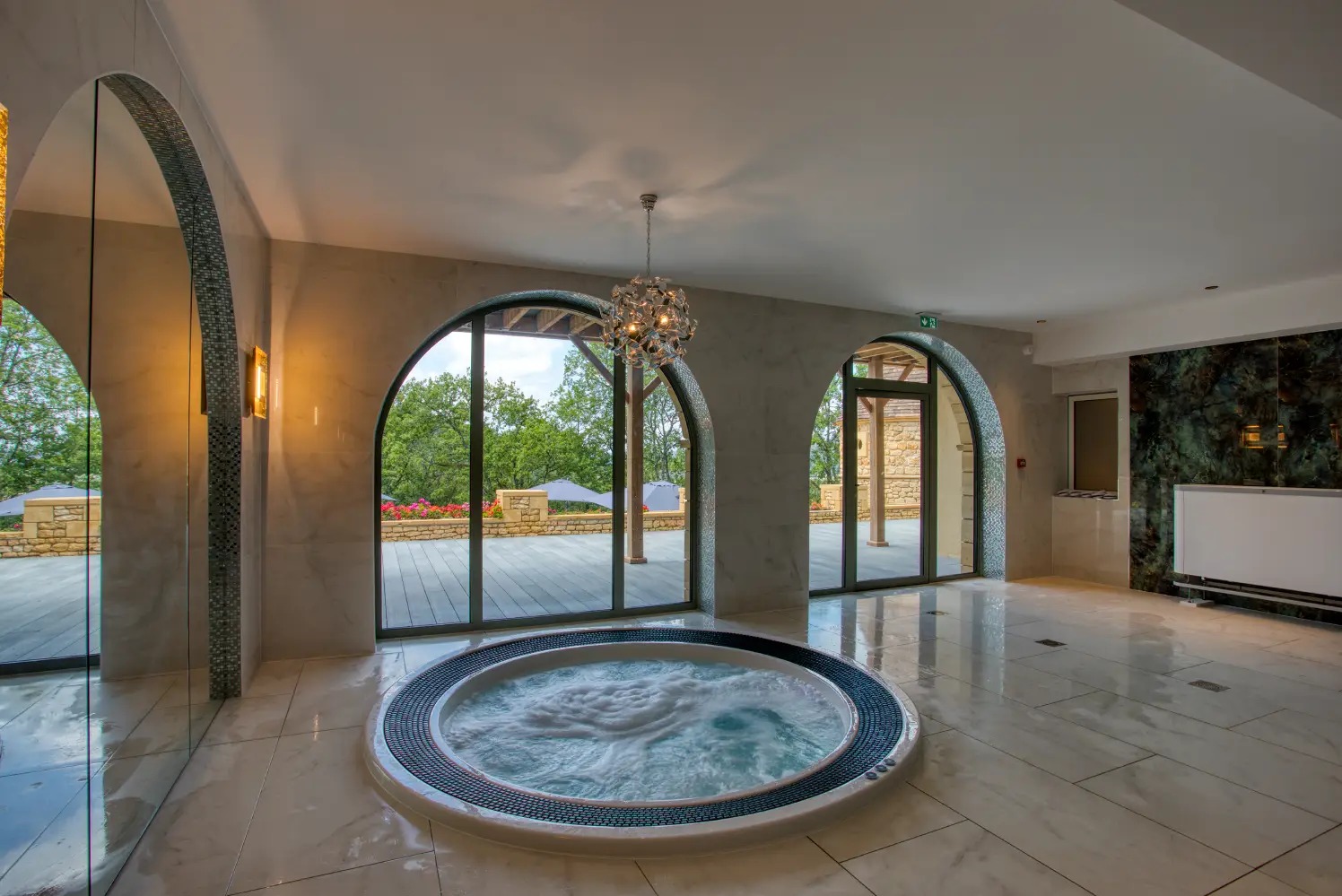 Ekskluzywne wnętrze z jacuzzi i przeszklonymi, łukowymi wyjściami na taras. Marmurowe płytki, dekoracyjne oświetlenie i widok na zieleń.