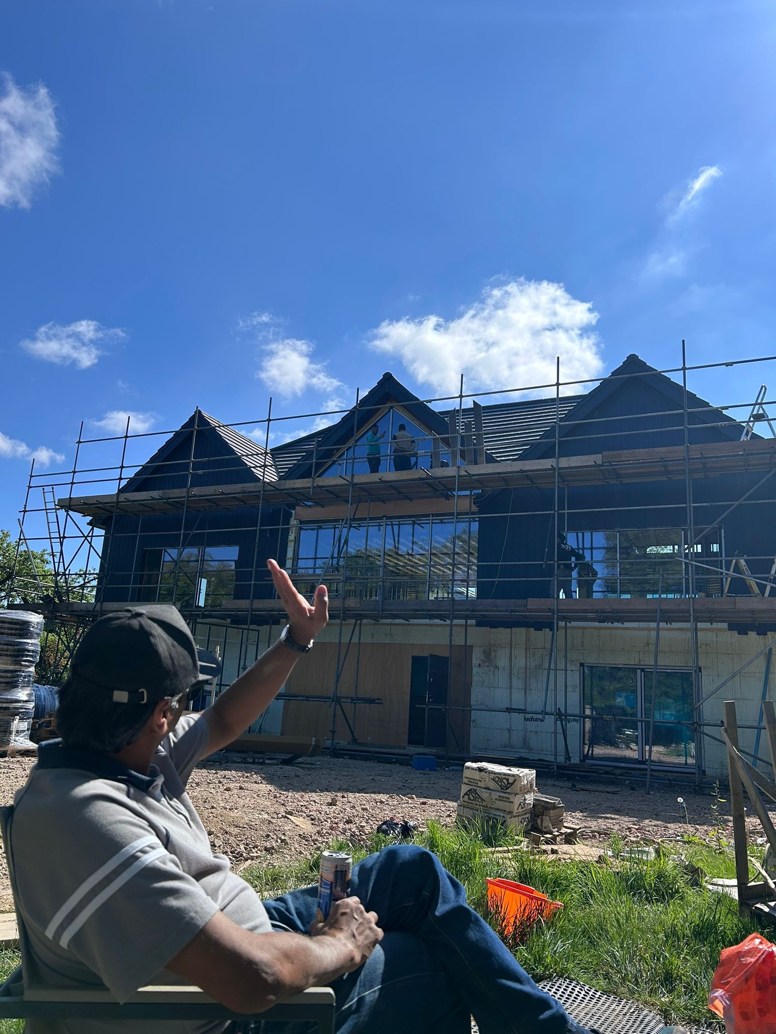 Dom w trakcie budowy z rusztowaniem, widoczne okna i robotnicy na górze, mężczyzna siedzi na krześle z puszką w ręku i gestykuluje.