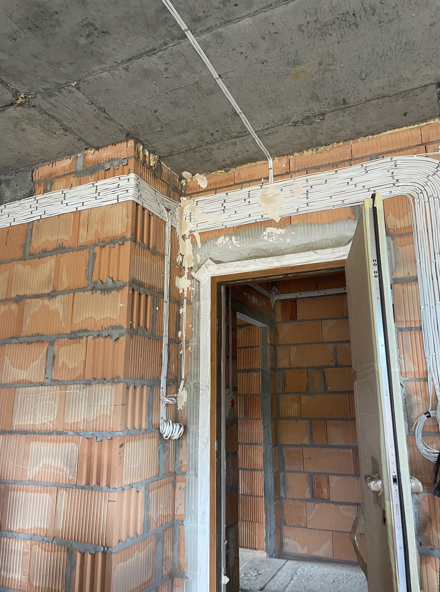 Surowy mur z instalacją elektryczną w białych listwach, widoczne przewody i puszki. Otwór drzwiowy z częściowo zamontowanymi drzwiami. Stan surowy.