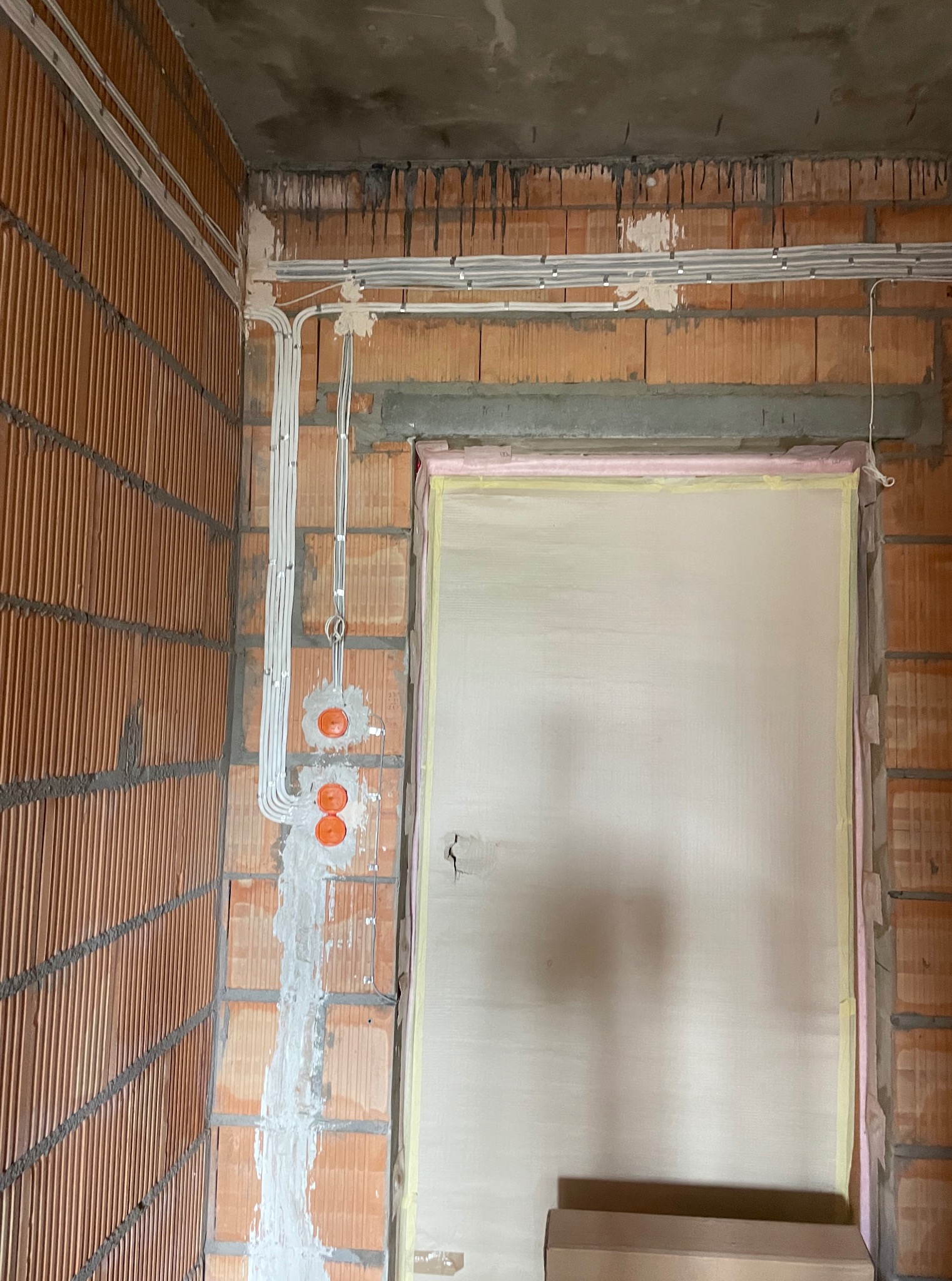 Instalacja elektryczna w trakcie montażu: kable prowadzone po ścianie z cegły, puszki elektryczne osadzone w ścianie, widoczne mocowania kabli. Surowy stan wykończenia.