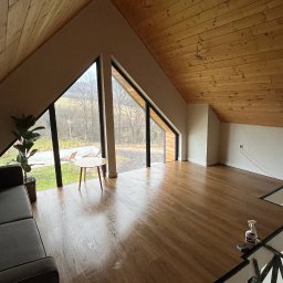 BUDUJ Z NAMI - Jasne poddasze z drewnianym sufitem i podłogą. Widoczne trójkątne okna z widokiem na zewnątrz, sofa i mały stolik. Podłoga błyszcząca, prawdopodobnie po czyszczeniu.