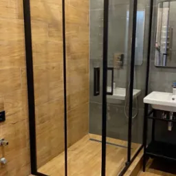 Nowoczesna kabina prysznicowa z czarnymi profilami i drewnopodobnymi płytkami, uzupełniona o umywalkę z szafką i lustro, tworzy spójną aranżację łazienki.