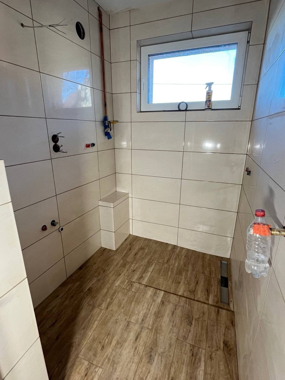 Wykończona łazienka z drewnopodobną podłogą i białymi płytkami. Widoczne przyłącza wodne, okno z chemią i plastikowa butelka. Nowoczesny minimalizm.