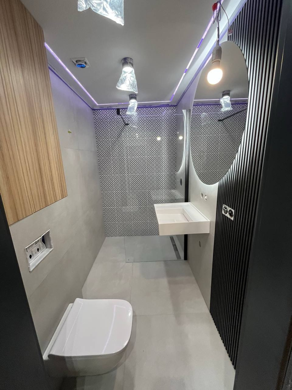 Nowoczesna łazienka po remoncie: biała armatura, geometryczne płytki na ścianie, okrągłe lustro z oświetleniem i drewniane akcenty. Minimalistyczny design.