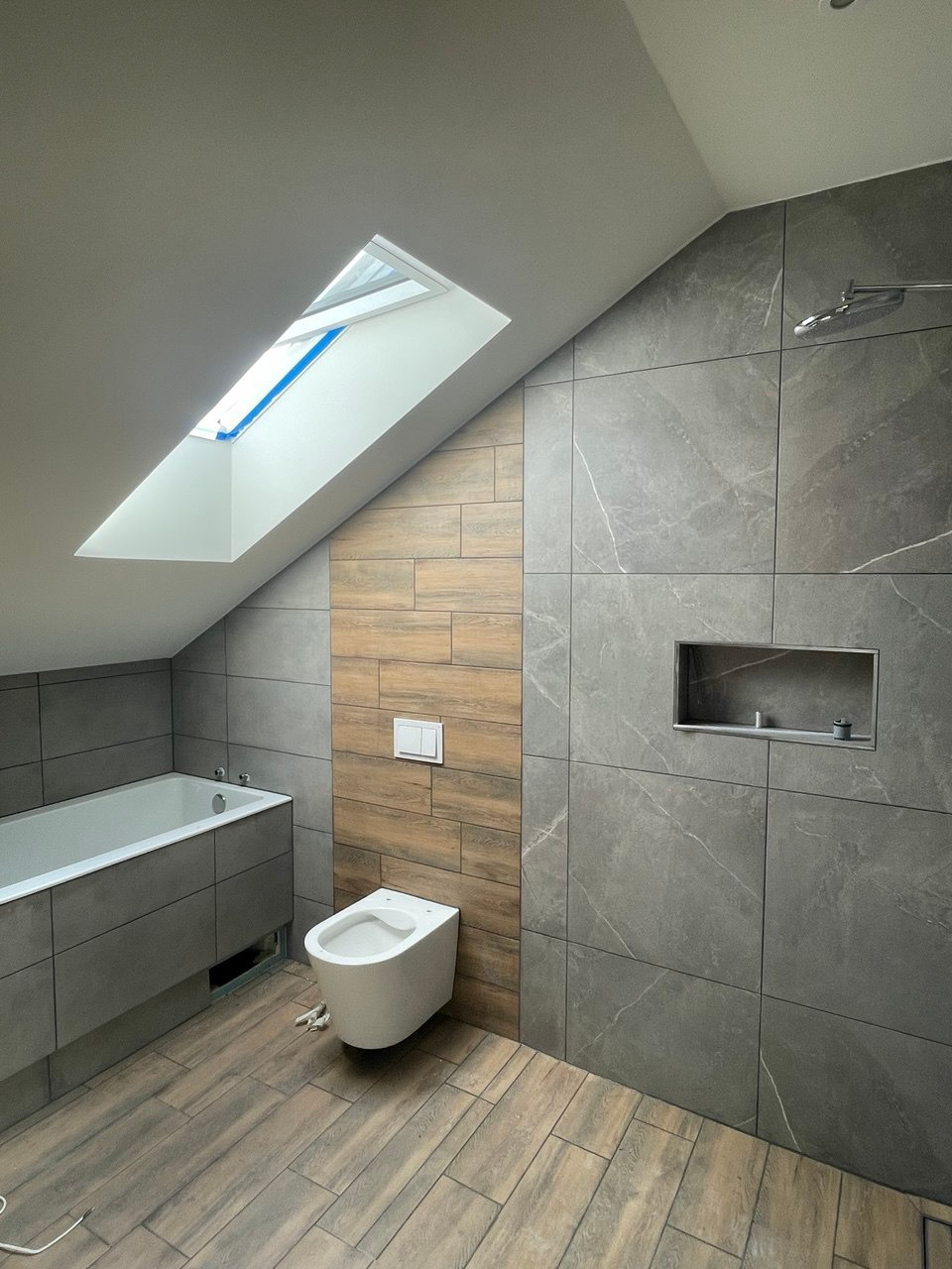 Nowoczesna łazienka na poddaszu z drewnopodobną podłogą i szarymi płytkami imitującymi marmur, akcentowana drewnianą ścianką z toaletą pod oknem dachowym.