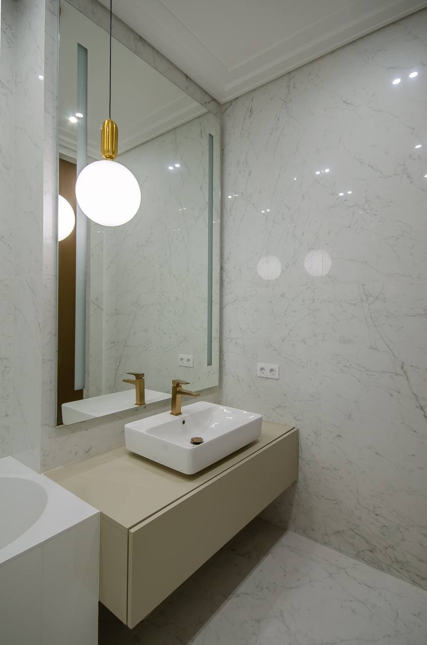 Elegancka łazienka z marmurowymi płytkami, złota armatura i okrągła lampa. Umywalka nablatowa na szafce w kolorze beżowym. Nowoczesny design.