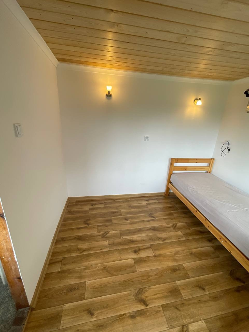 Pokój z drewnianą podłogą, boazerią na suficie i łóżkiem. Dwa kinkiety na ścianie oświetlają wnętrze. Widok na świeżo wykończone wnętrze.