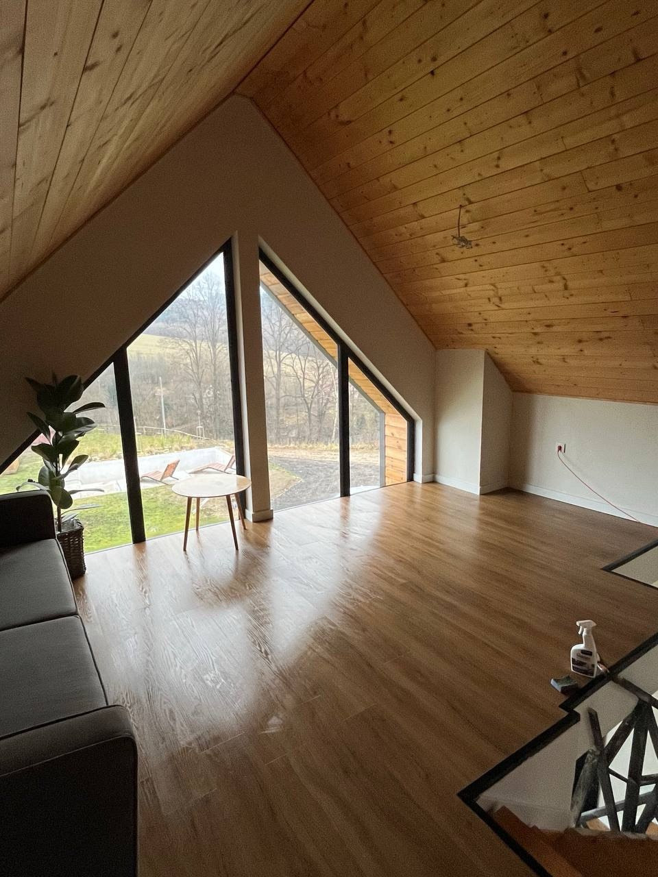Jasne poddasze z drewnianym sufitem i podłogą. Widoczne trójkątne okna z widokiem na zewnątrz, sofa i mały stolik. Podłoga błyszcząca, prawdopodobnie po czyszczeniu.