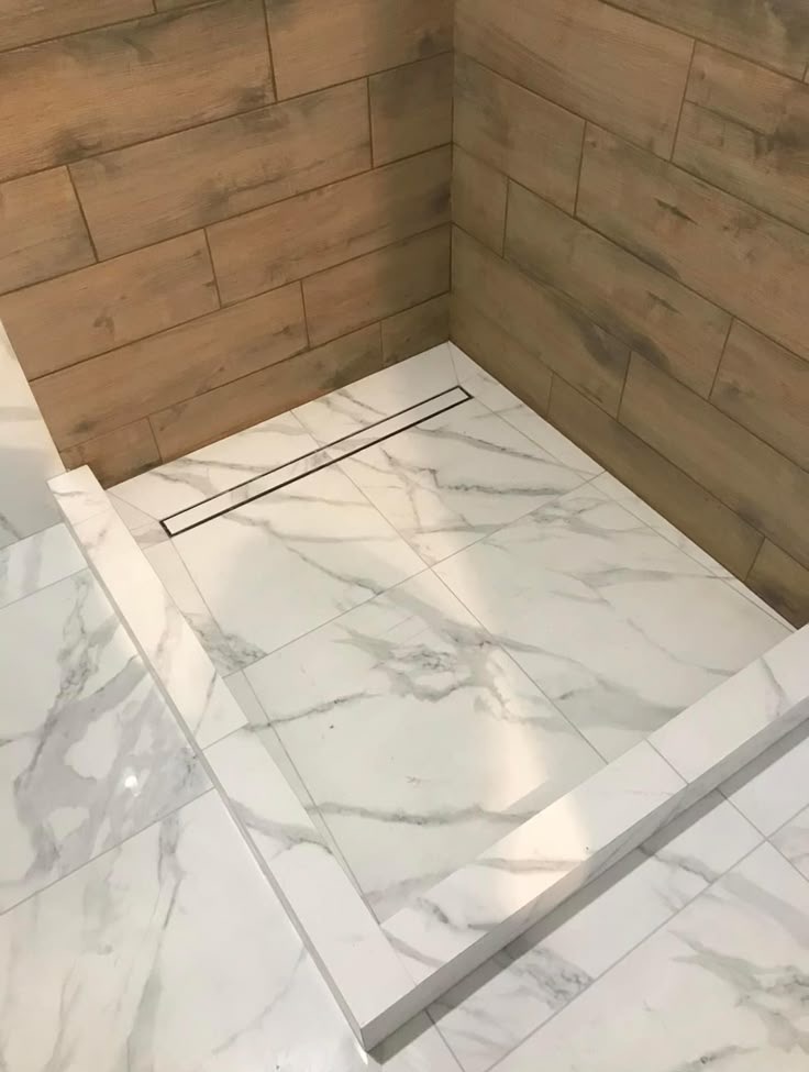 Wykończony brodzik z odpływem liniowym, wyłożony białym marmurem. Ściany wyłożone płytkami imitującymi drewno. Minimalistyczny design, czysty kadr.