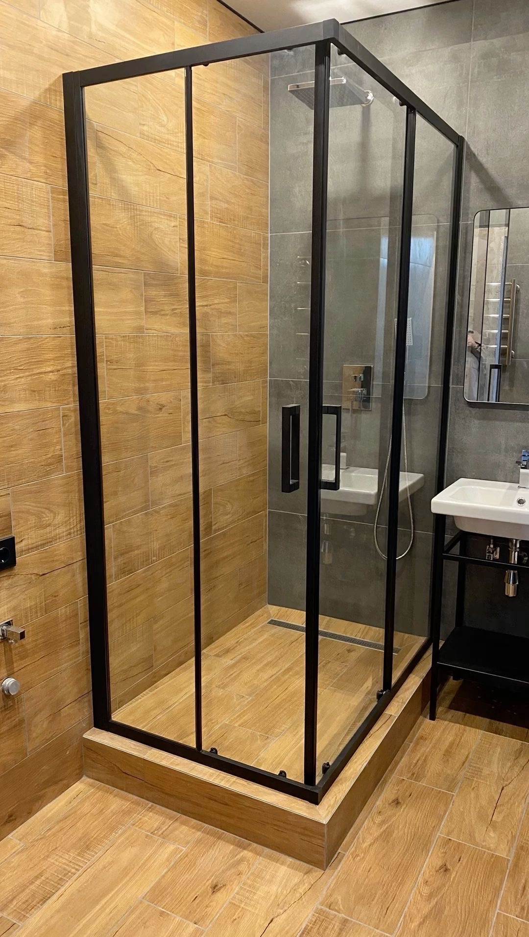 Nowoczesna kabina prysznicowa z czarnymi profilami i drewnopodobnymi płytkami, uzupełniona o umywalkę z szafką i lustro, tworzy spójną aranżację łazienki.