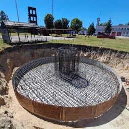 WAMKO Mateusz Kowalczyk - Okrągły wykop zbrojony siatką i pionowymi prętami, przygotowany do zalania betonem, Latowicz. Widok z góry, błękitne niebo, budynek w tle.