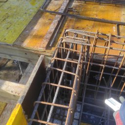 WAMKO Mateusz Kowalczyk - Zbrojenie fundamentów na żółtym szalunku, widoczne fragmenty butów robotników. Detal zbrojenia z bliska, ukazujący etap prac budowlanych.