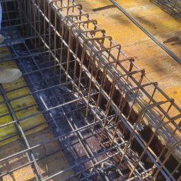 WAMKO Mateusz Kowalczyk - Zbrojenie fundamentów na żółtym szalunku, widoczne buty pracownika. Konstrukcja z prętów stalowych przygotowana do zalania betonem. Detal budowy z bliska.
