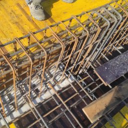 WAMKO Mateusz Kowalczyk - Zbrojenie fundamentów z żółtą szalunką, widok z góry. Stalowe pręty tworzą siatkę, częściowo zardzewiałe. Fragment buta roboczego w rogu kadru.