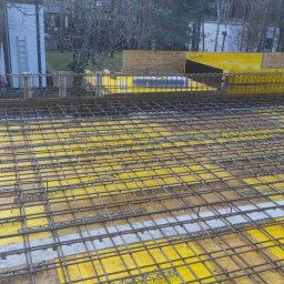 WAMKO Mateusz Kowalczyk - Zbrojenie stropu na żółtym szalunku, widoczna siatka z prętów stalowych. W tle pracownik w odblaskowej kamizelce i budynek. Ujęcie z góry, podkreślające regularność siatki.