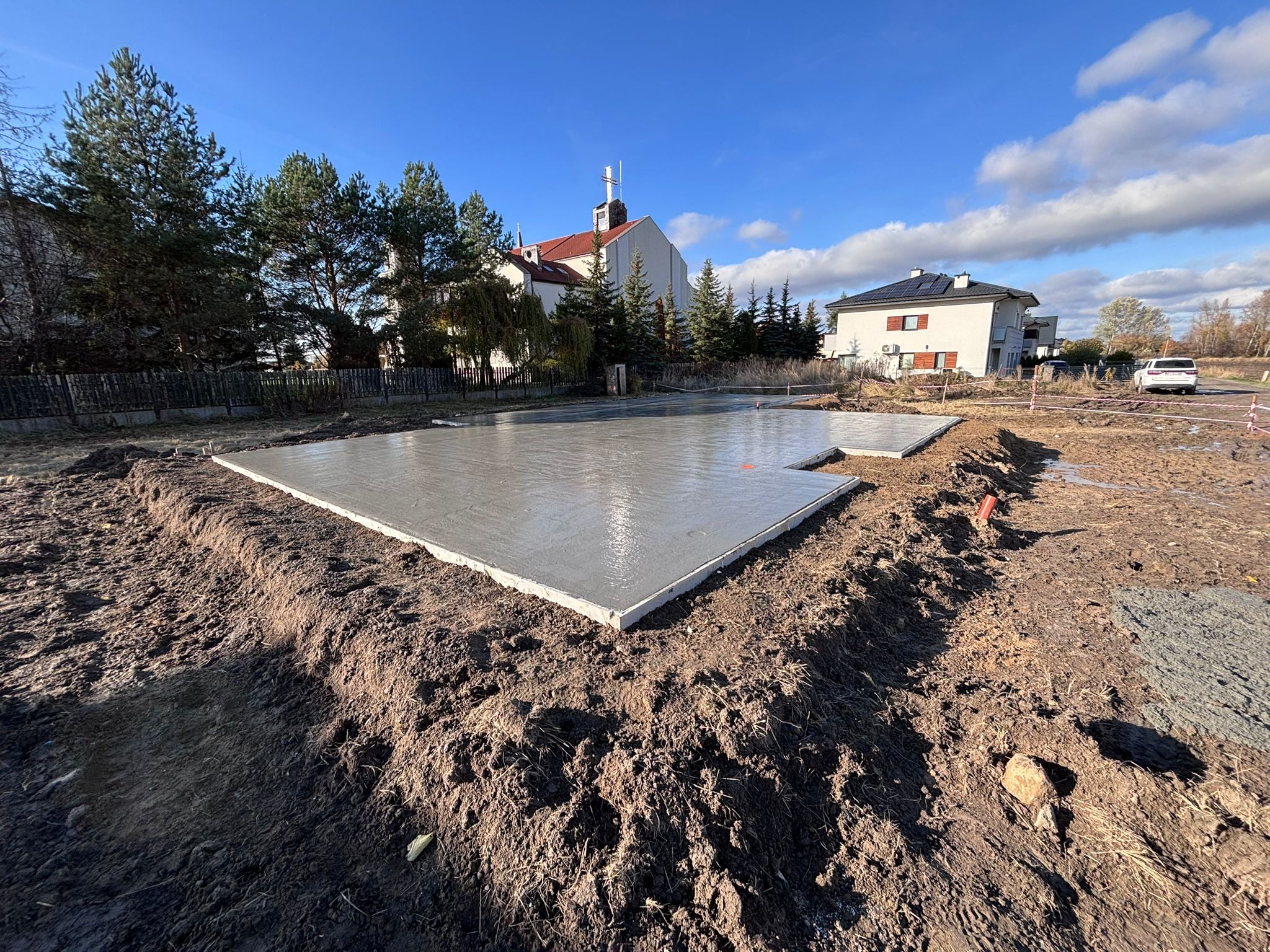 Wylana, świeża płyta fundamentowa na budowie domu jednorodzinnego w słoneczny dzień. Widoczne wykopane fundamenty i kościół w tle.