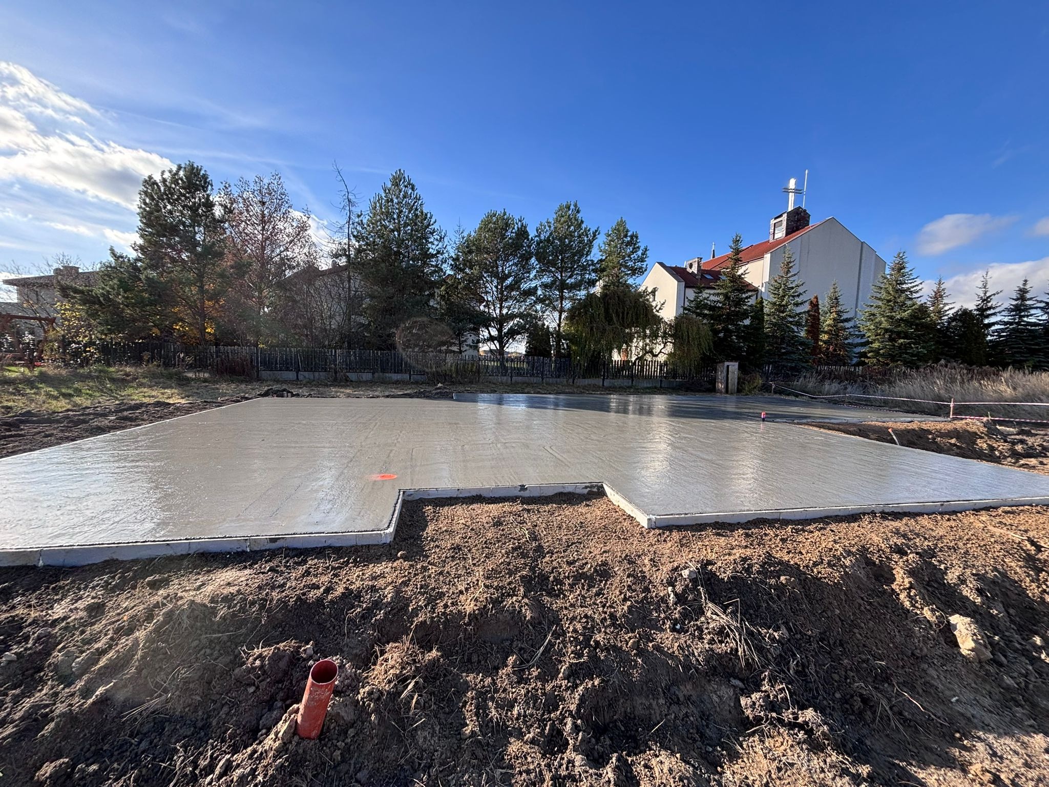 Świeżo wylana, gładka płyta fundamentowa z widocznymi obrzeżami, na tle błękitnego nieba i zabudowań. Widoczny fragment rury w ziemi.