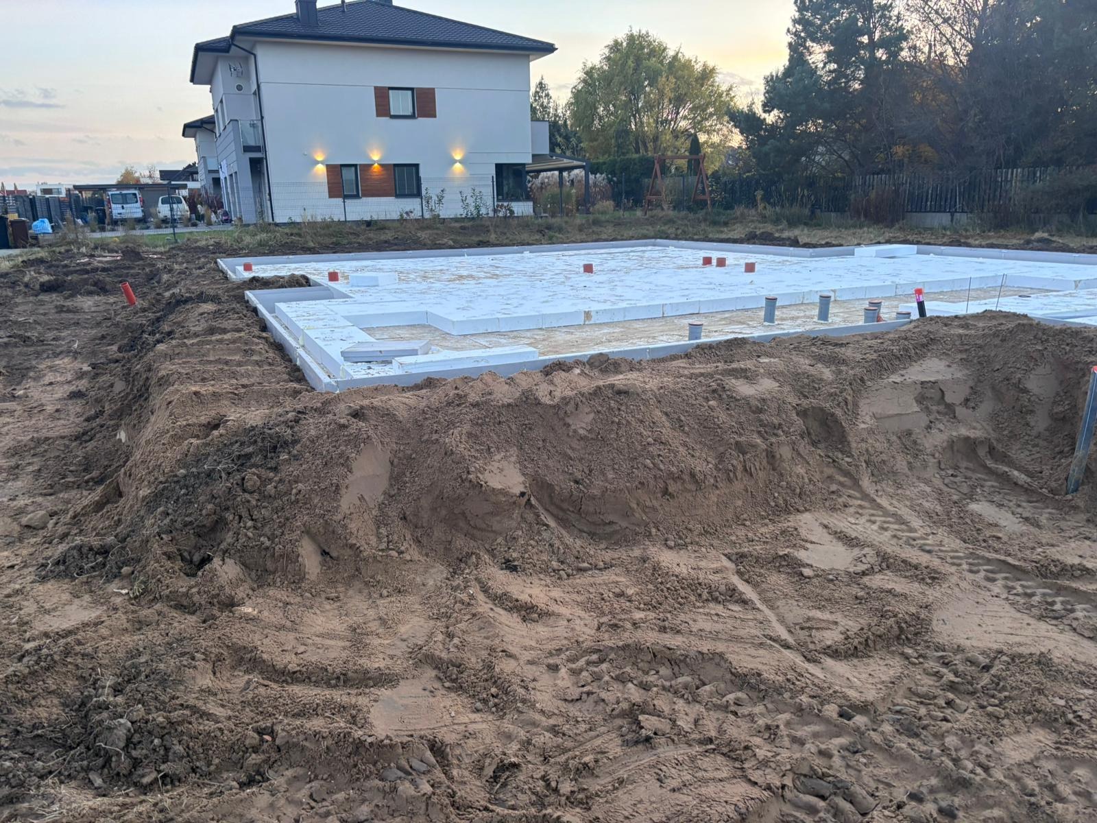 Fundamenty domu jednorodzinnego w trakcie budowy, z widoczną izolacją termiczną i instalacjami. W tle dom w stanie surowym zamkniętym i drzewa.