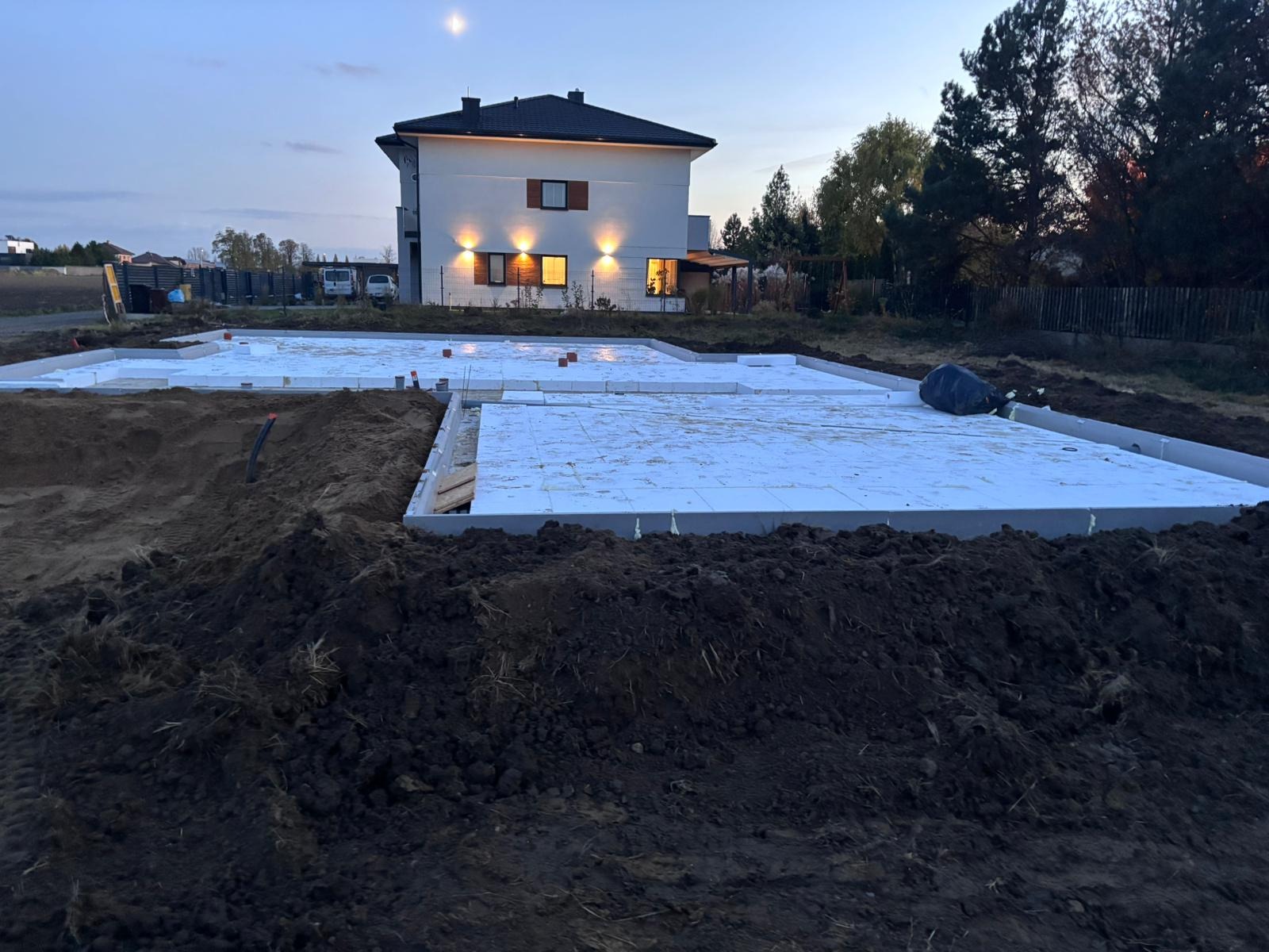 Fundamenty domu jednorodzinnego z izolacją termiczną, widoczne oświetlone okna budynku w tle, wieczorny krajobraz z księżycem w górze.