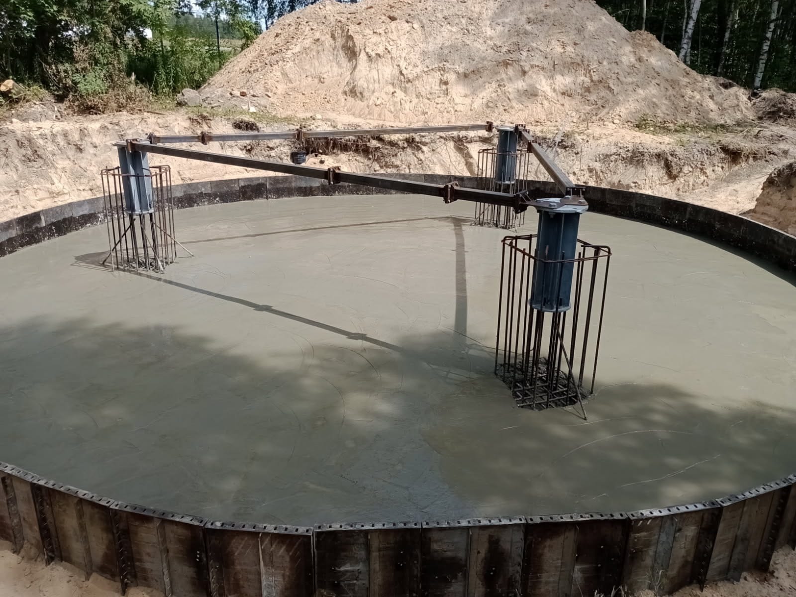 Zalany fundament okrągły z metalowym szalunkiem i zbrojeniem, widok z góry. W tle ziemia i drzewa.