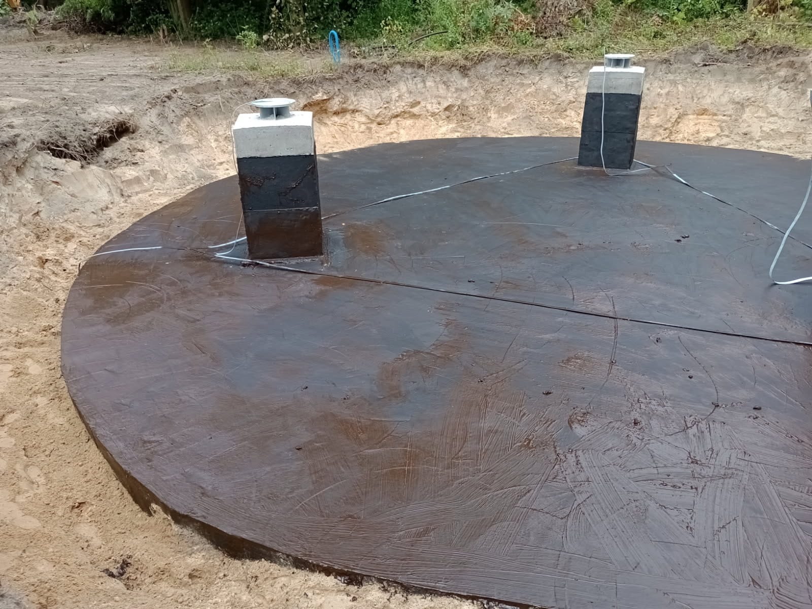 Zabezpieczona, okrągła płyta fundamentowa w wykopie, z dwoma pionowymi kominami wentylacyjnymi. Widoczne ślady pędzla na powierzchni płyty i piasek w wykopie.