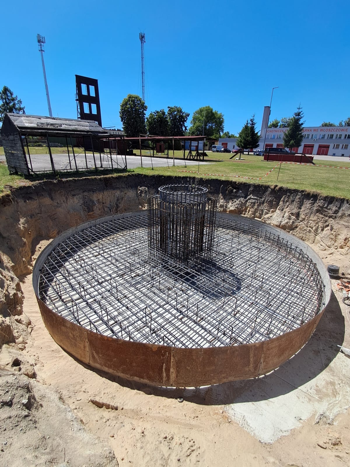 Okrągły wykop zbrojony siatką i pionowymi prętami, przygotowany do zalania betonem, Latowicz. Widok z góry, błękitne niebo, budynek w tle.