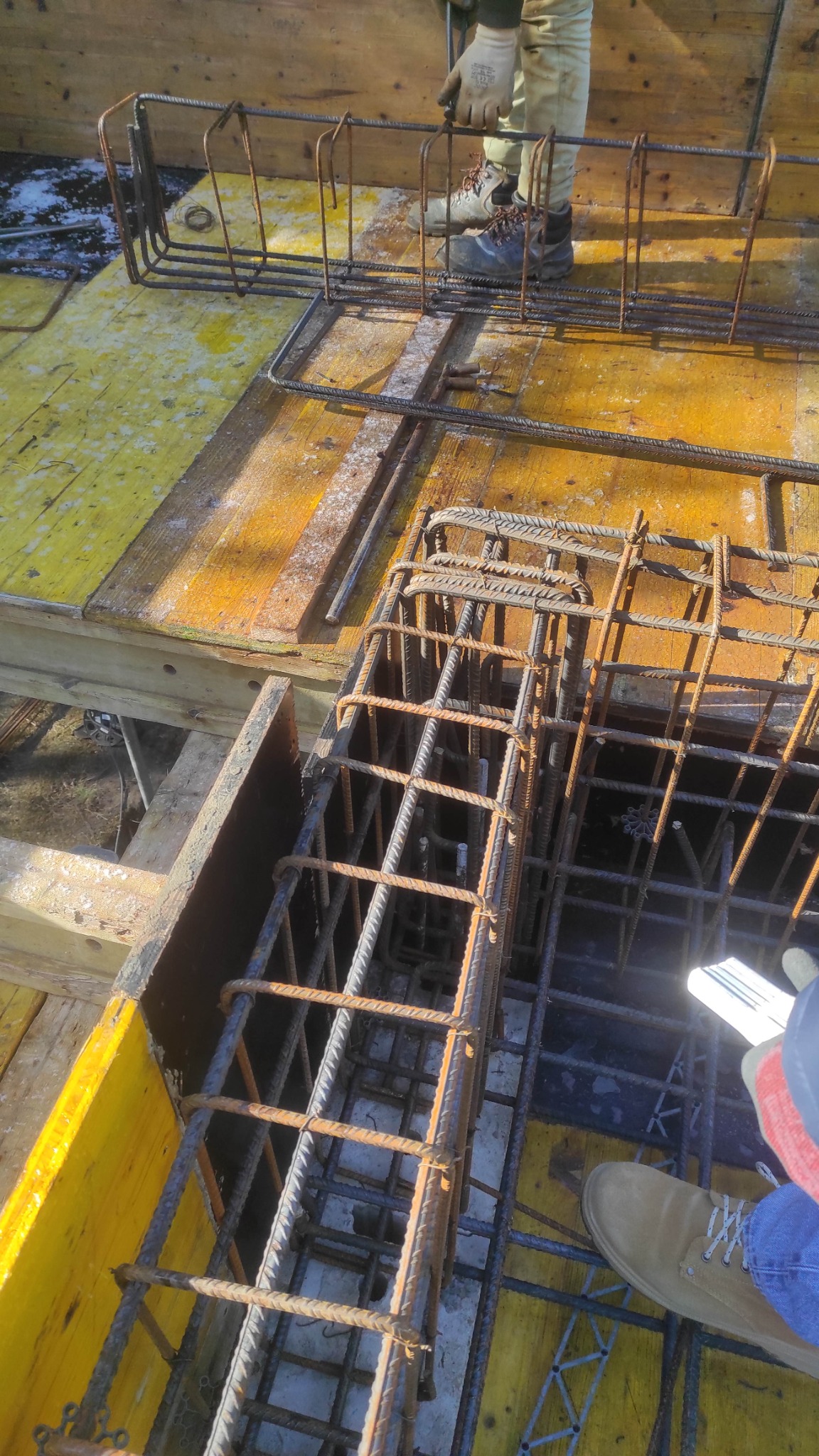 Zbrojenie fundamentów na żółtym szalunku, widoczne fragmenty butów robotników. Detal zbrojenia z bliska, ukazujący etap prac budowlanych.