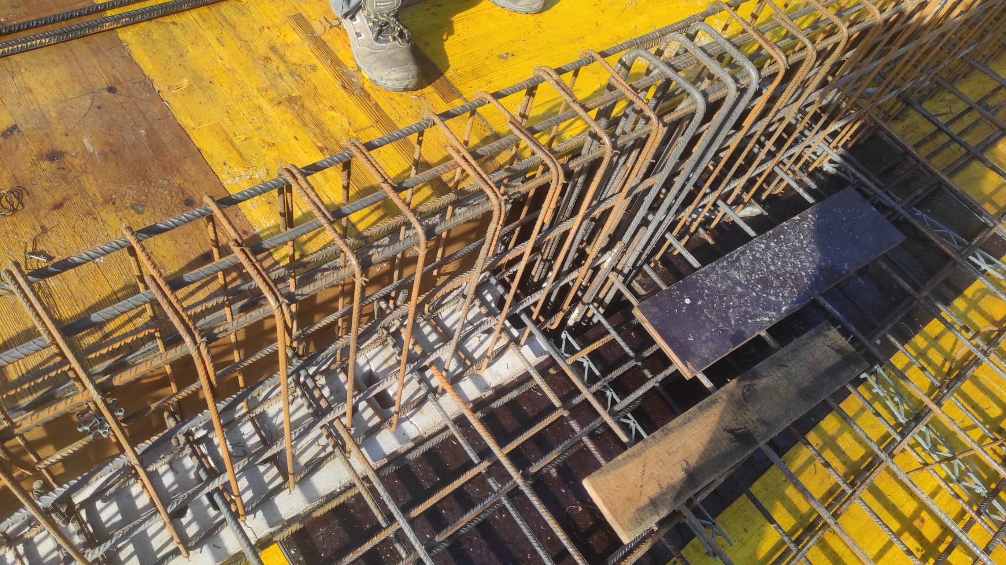 Zbrojenie fundamentów z żółtą szalunką, widok z góry. Stalowe pręty tworzą siatkę, częściowo zardzewiałe. Fragment buta roboczego w rogu kadru.