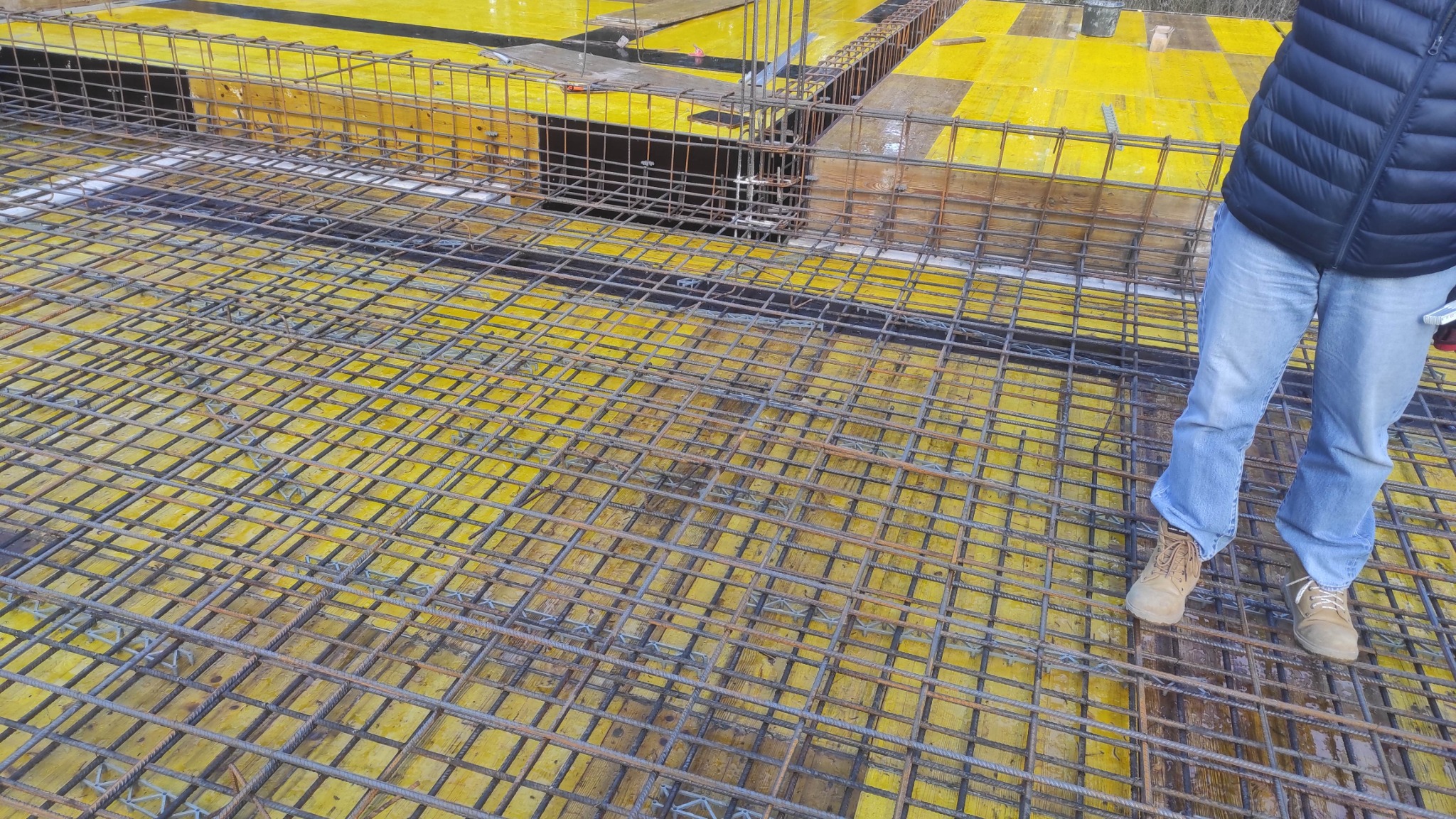 Zbrojenie stropu na żółtym szalunku, widok z góry. Fragment sylwetki osoby w jeansach i roboczych butach. Detal budowy z bliska, ukazujący siatkę zbrojeniową.