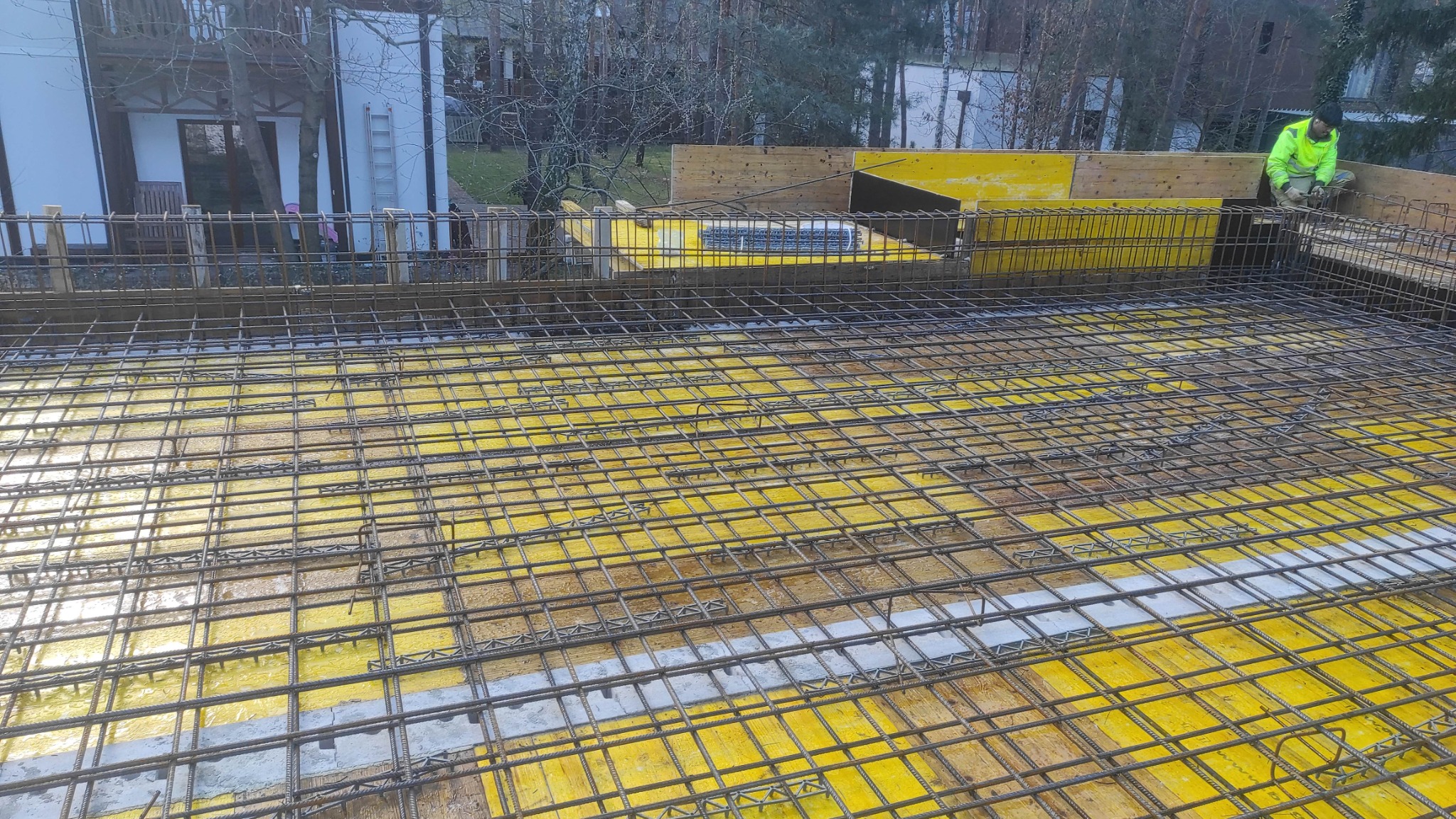 Zbrojenie stropu na żółtym szalunku, widoczna siatka z prętów stalowych. W tle pracownik w odblaskowej kamizelce i budynek. Ujęcie z góry, podkreślające regularność siatki.
