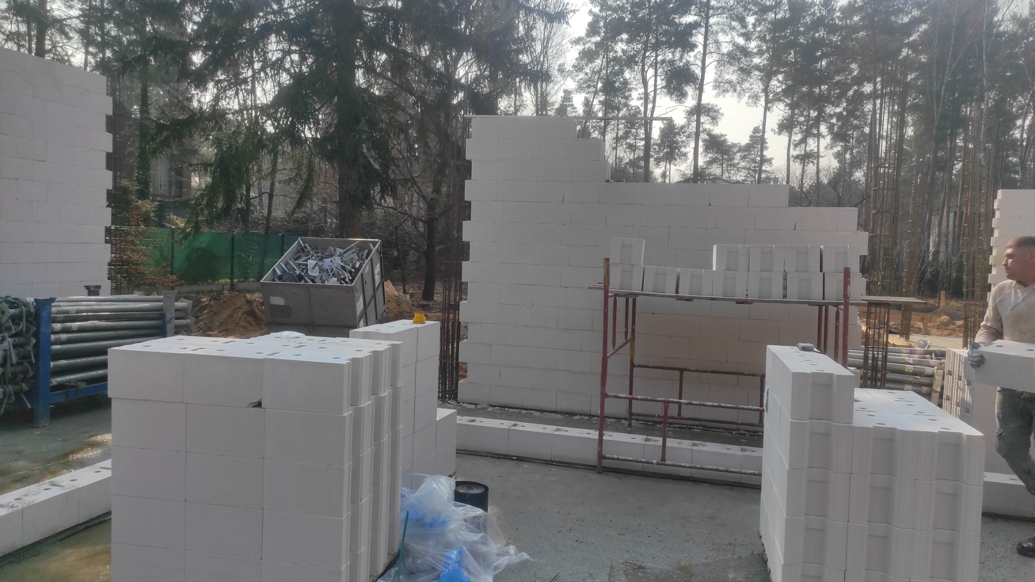 Wznoszenie ścian z białych bloczków na budowie domu jednorodzinnego. Widoczne rusztowania, materiały budowlane i pracownik w trakcie pracy. Tło stanowią drzewa.