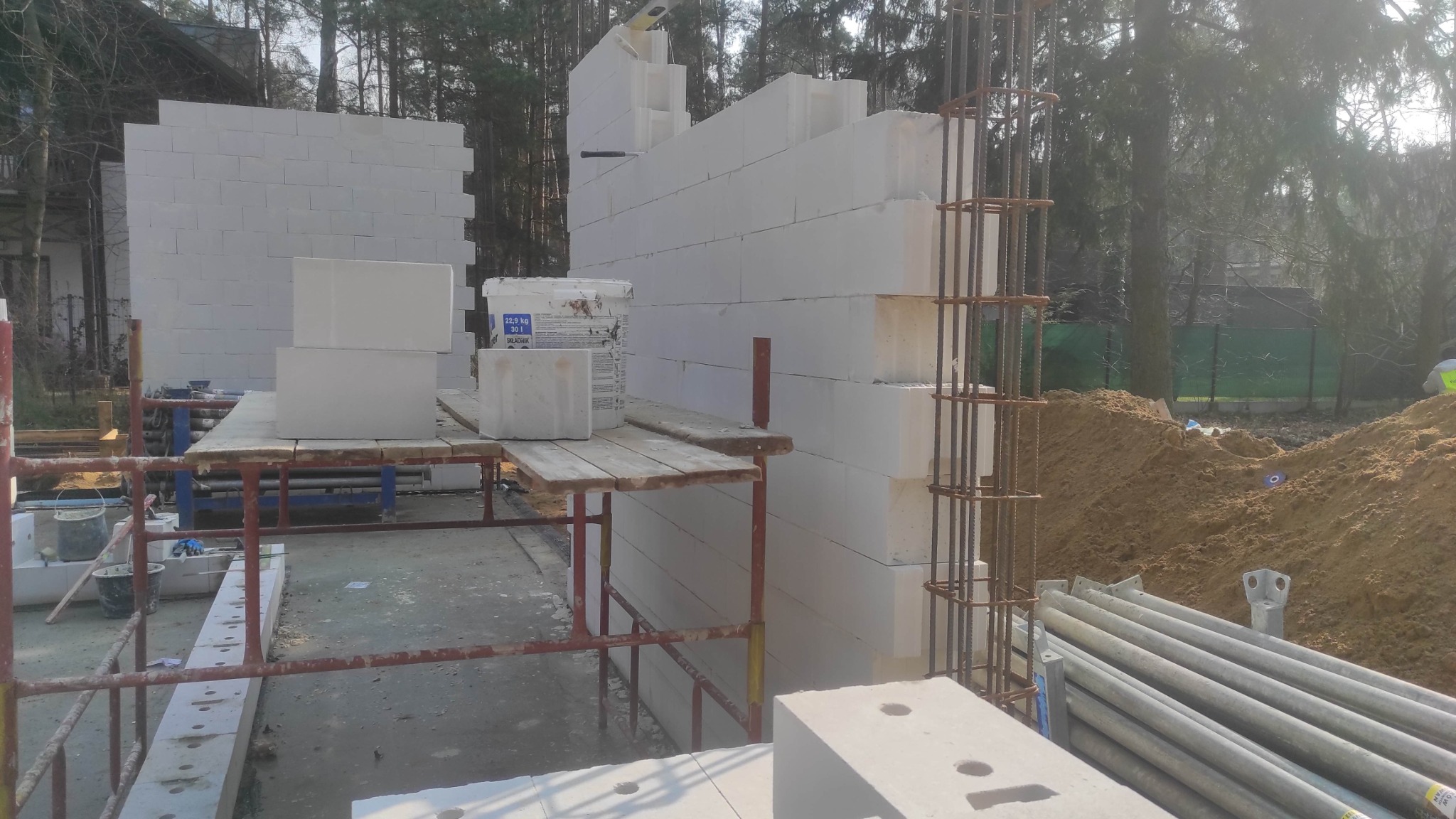 Budowa ściany z białych bloczków, widoczne zbrojenia i rusztowania. Prace murarskie w toku, na zewnątrz, w tle drzewa i częściowo widoczny budynek.