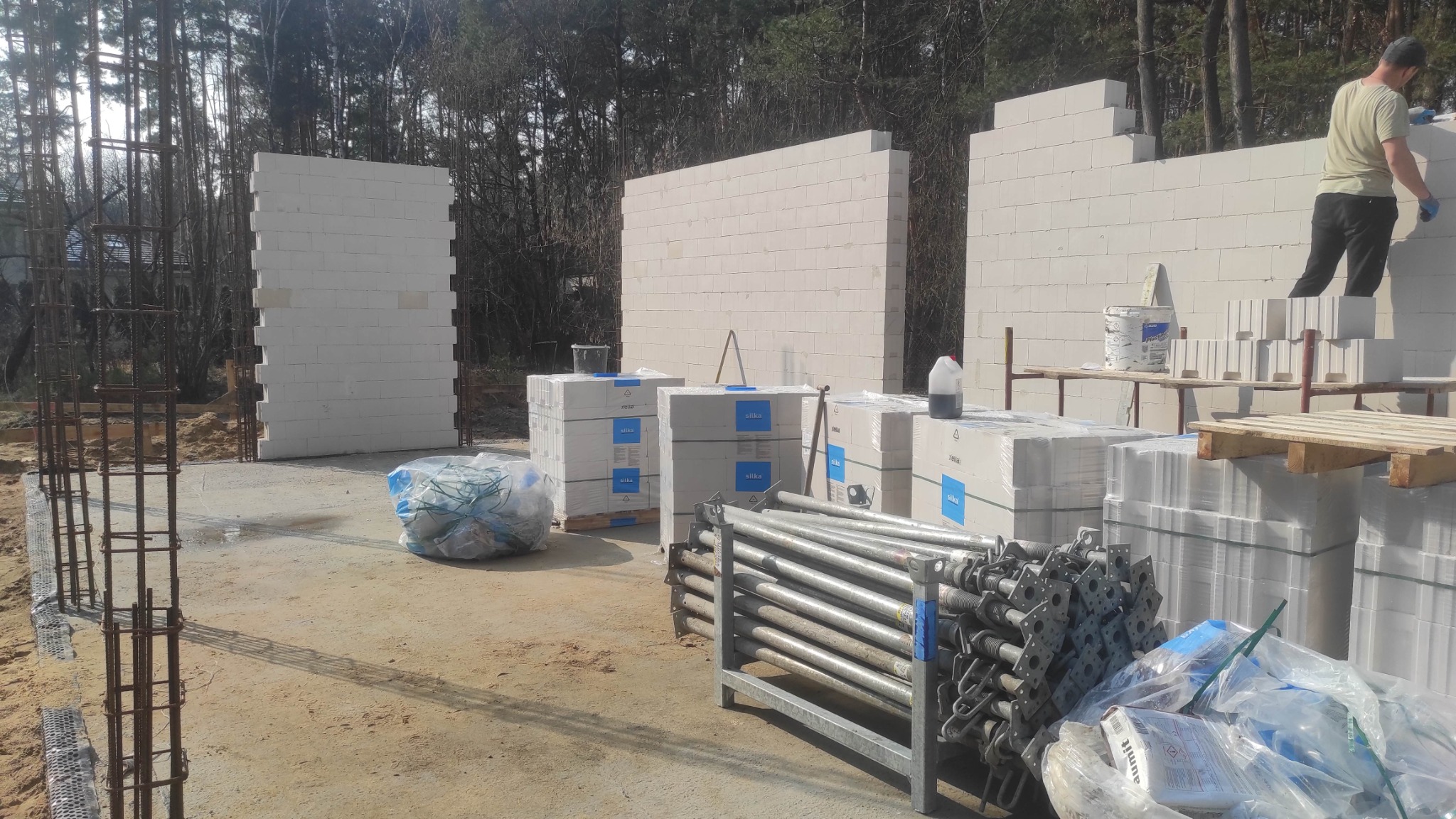 Budowa domu z bloczków Silka: wzniesione ściany, zbrojenie fundamentów, palety z materiałami budowlanymi i stemple budowlane na placu budowy w słoneczny dzień.