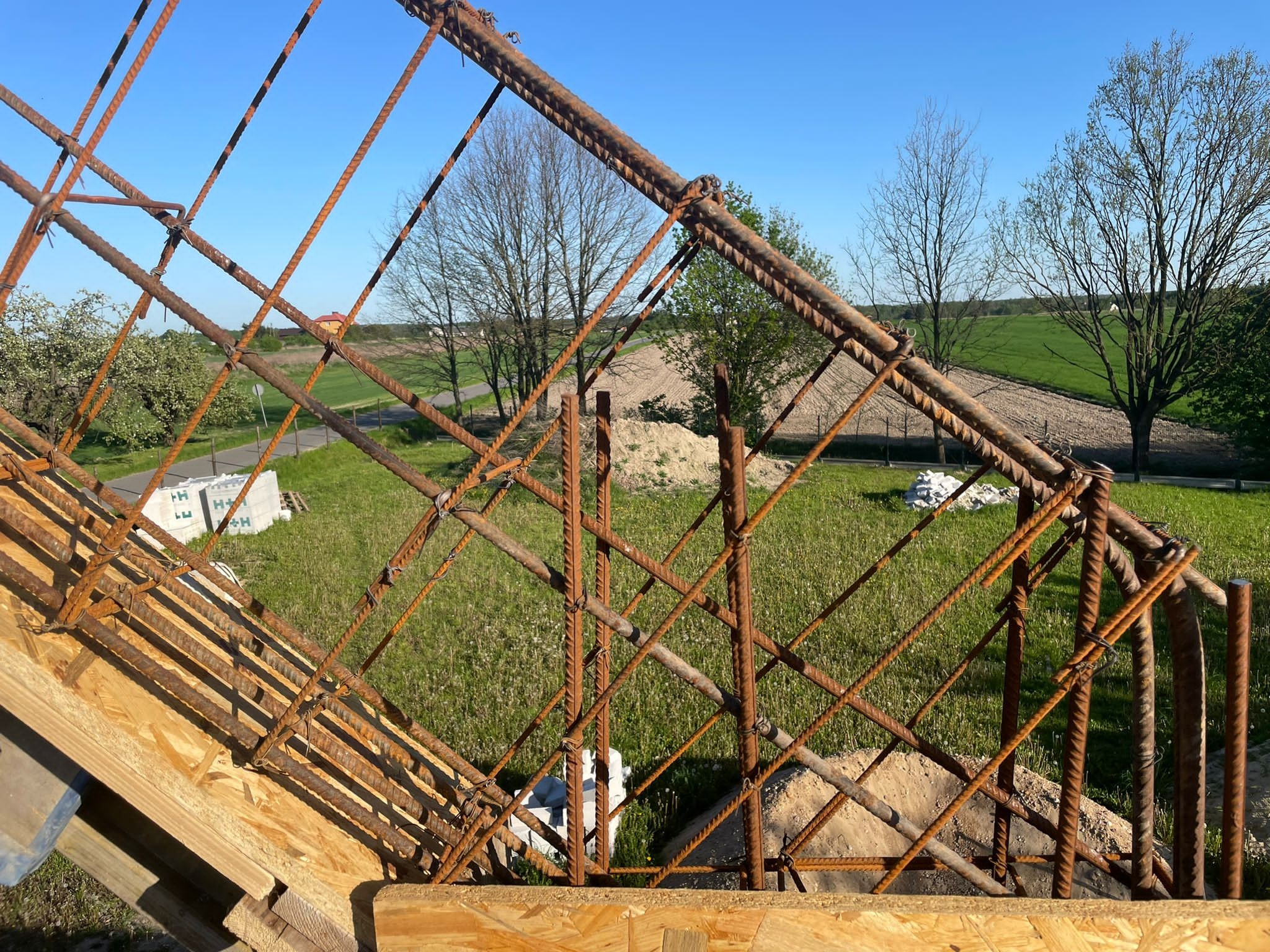 Zbrojenie konstrukcji żelbetowej na budowie, widok z góry na siatkę z prętów stalowych, w tle zielone pola i błękitne niebo. Elementy szalunku z płyty OSB.