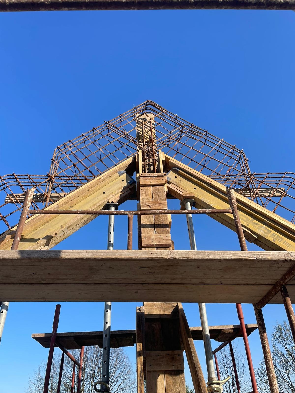 Zbrojenie dachu domu jednorodzinnego z drewnianym szalunkiem, widok z dołu na tle błękitnego nieba. Konstrukcja w trakcie budowy, widoczne elementy rusztowania.