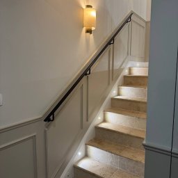 Renovest - Wykończenie Wnętrz - Schody z beżowego kamienia z podświetleniem LED, czarna poręcz, ściany z dekoracyjnymi panelami i kinkietami. Klasyczny styl wykończenia wnętrza.